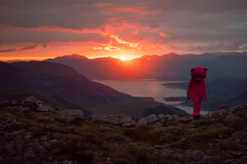 LUAP artist Paul Robinson dressed in his pink bear suit atop a mountain with a sunset behind him